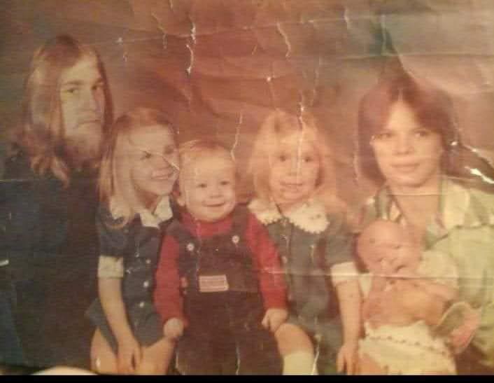 A gathering of five children and two adults dressed in 1970s attire, smiling and posing together.