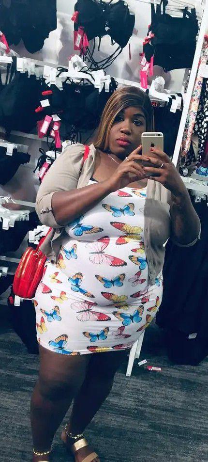 A woman takes a selfie in a vibrant dress adorned with butterflies in a retail store.