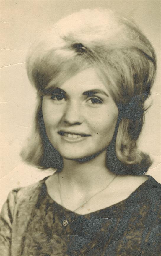 A young woman smiles at the camera, wearing a top and jewelry. Her light hair frames her face.