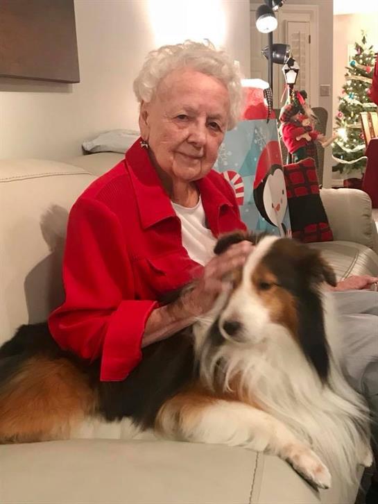 An elderly woman pets a dog while sitting on a sofa in a festive living room with decorations.