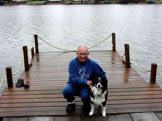 Man sits on a dock with a dog by a lake. The sky is overcast, and water is still.