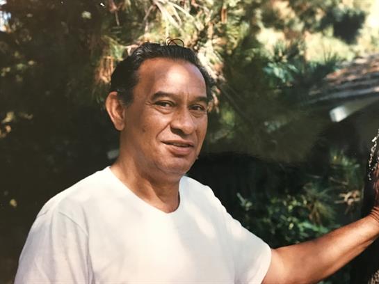 A man stands outdoors among the trees with a smile and a welcoming gesture towards the camera.