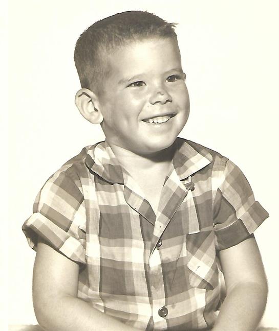 A young boy sits with a big smile in a checkered shirt. The background is bright and simple.