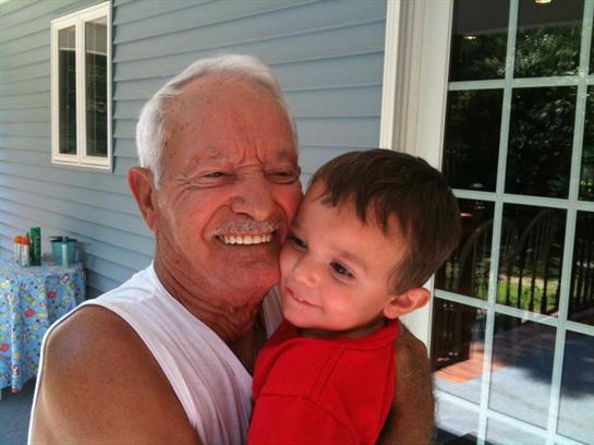An elderly man smiles while holding a young boy outside a house. They look happy together.