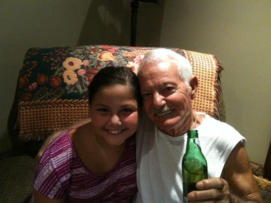 A young girl and an older man sit on a couch smiling while holding a drink in a cozy room.