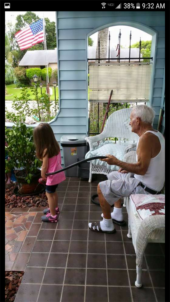 An elderly man holds a garden hose while a young girl waters plants. They enjoy gardening outside.