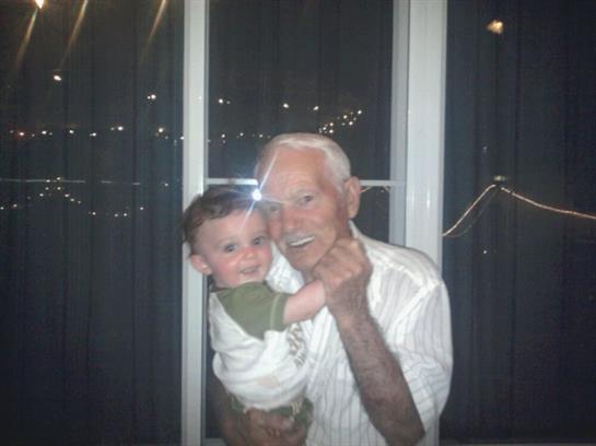 A grandfather smiles as he holds his grandchild close while standing by a lit window.
