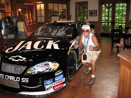 A man is smiling and posing near a black race car in a room with wood decor and other visitors.
