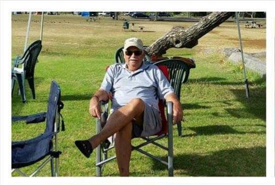 An elderly man relaxes in a chair on a grassy area under a tree while others are nearby.