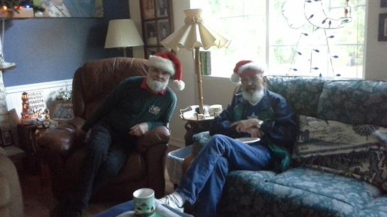 Two men are sitting in a living room. They are wearing Santa hats and smiling.