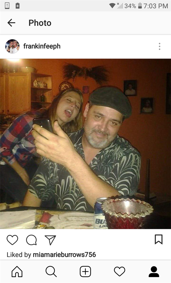 A man sits with a phone in hand while a woman sings playfully behind him during a gathering.