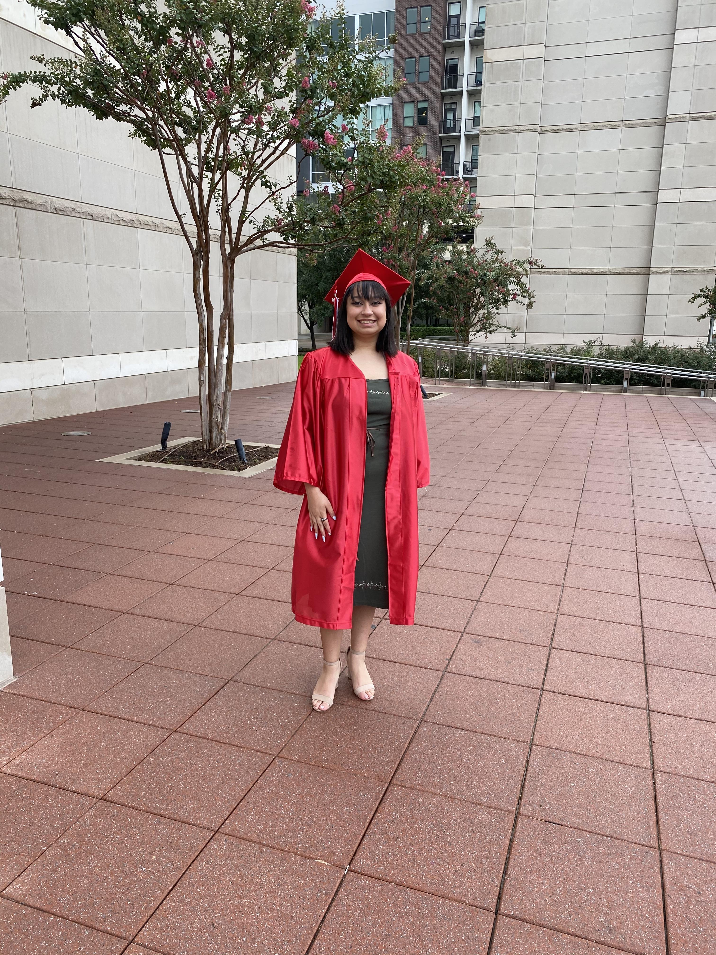 Person in a red cap and gown stands proudly on a paved area near urban buildings.