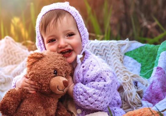 A happy toddler enjoys a sunny day while hugging a teddy bear and sitting on a cozy blanket.