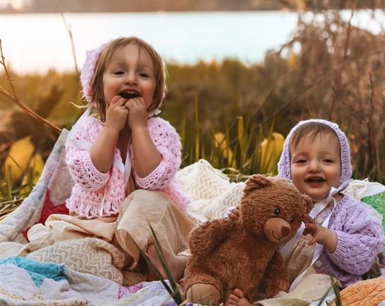 Two young girls dressed in cozy outfits are happily playing with a teddy bear beside a lake.