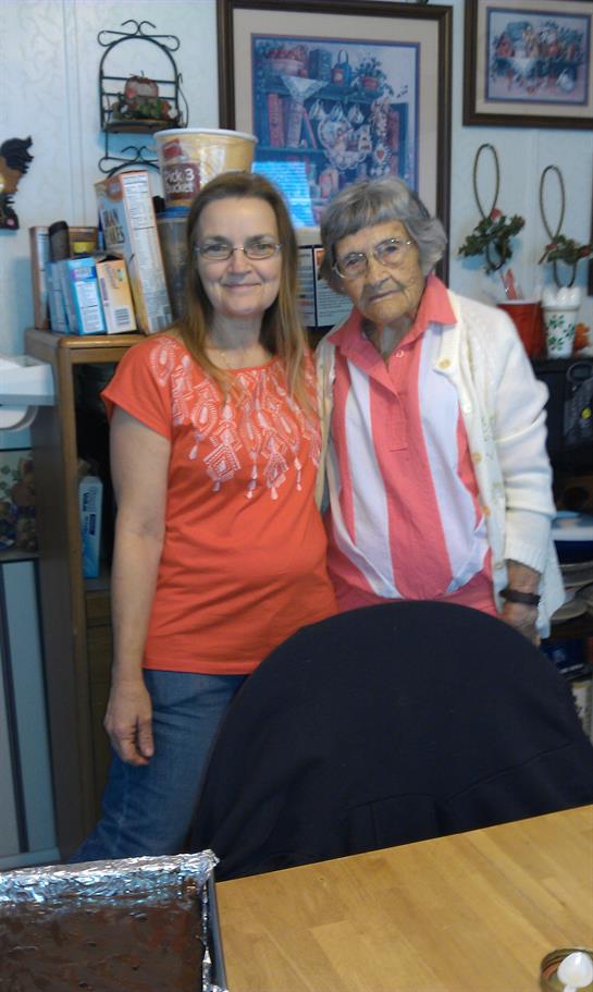Two smiling women stand close together in a cozy home with shelves around them.