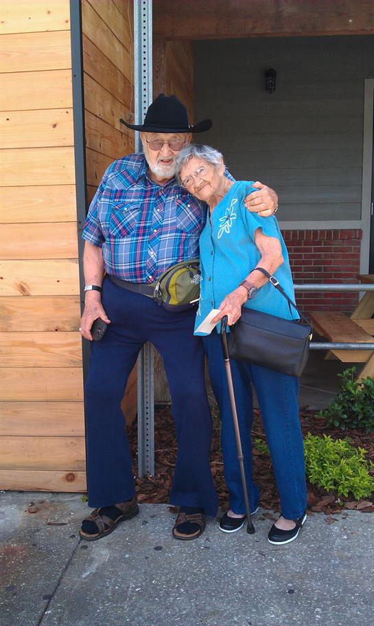 Older man and woman pose together outside a wooden structure with a warm smile and close embrace.