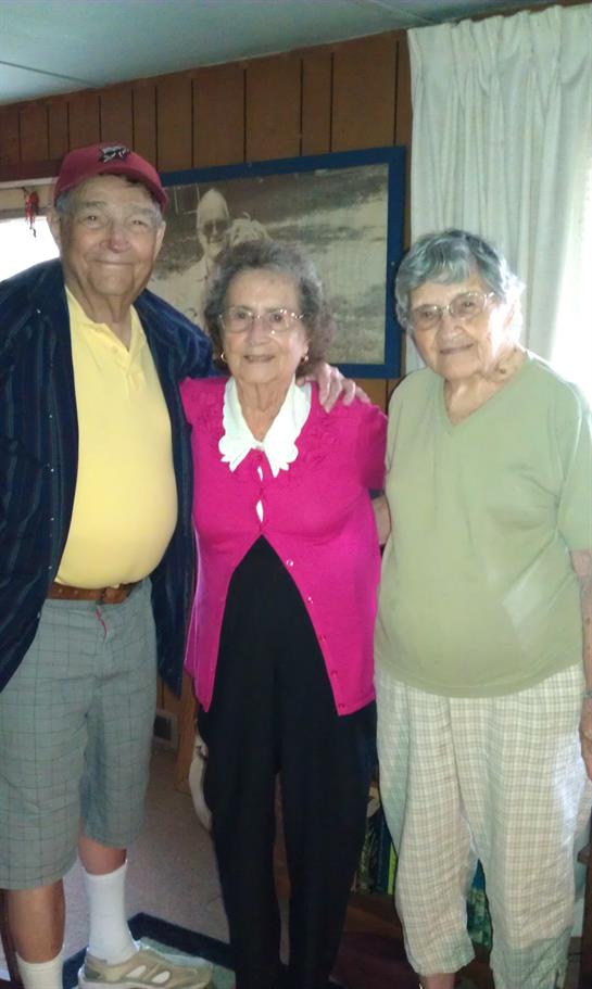 Three older adults stand together in a room with smiles. They pose closely, showing friendship.