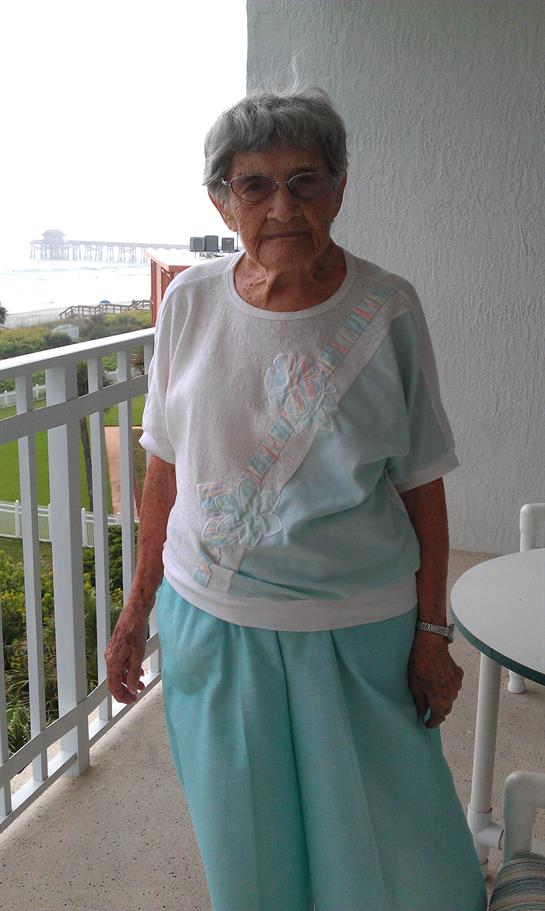 An elderly woman stands on a balcony looking at the water on a clear day near a bridge.