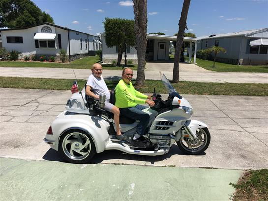 Two individuals are riding a custom motorcycle trike under bright sunshine in a quiet neighborhood.