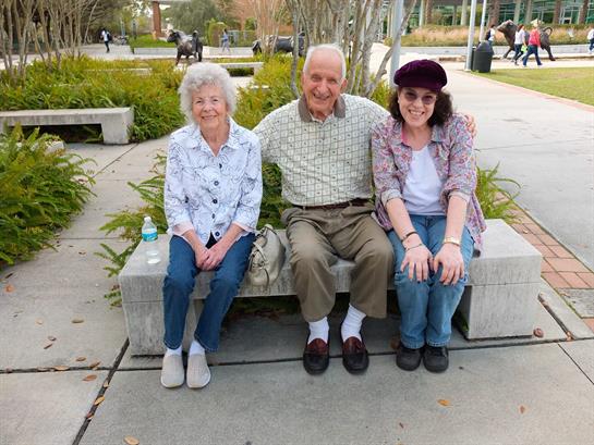 Three seniors share smiles while sitting on a bench in a lively urban park on a sunny day.