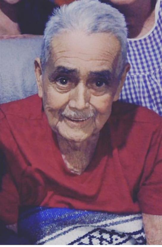 An elderly man smiles broadly while sitting in a comfortable chair at home during a family event.