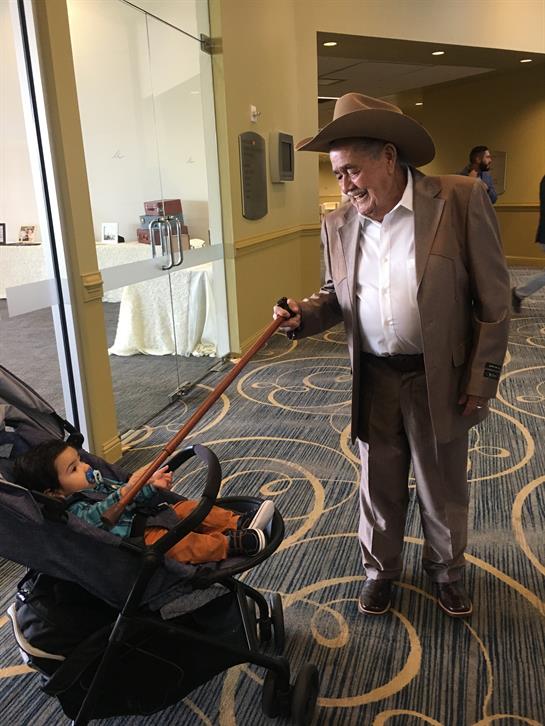 An elderly man wearing a cowboy hat smiles while engaging with a child in a stroller indoors.