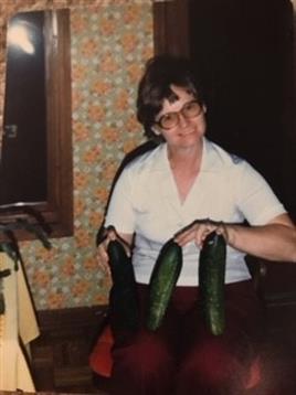 A woman poses with freshly picked cucumbers, showcasing her gardening success in a cozy kitchen.
