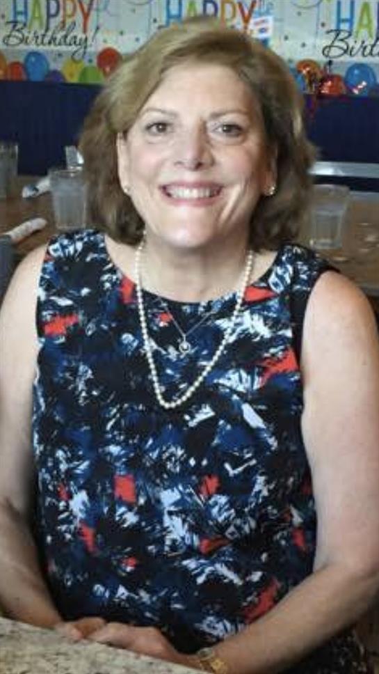 A cheerful woman in a floral dress and pearl necklace enjoys a casual event with friends.
