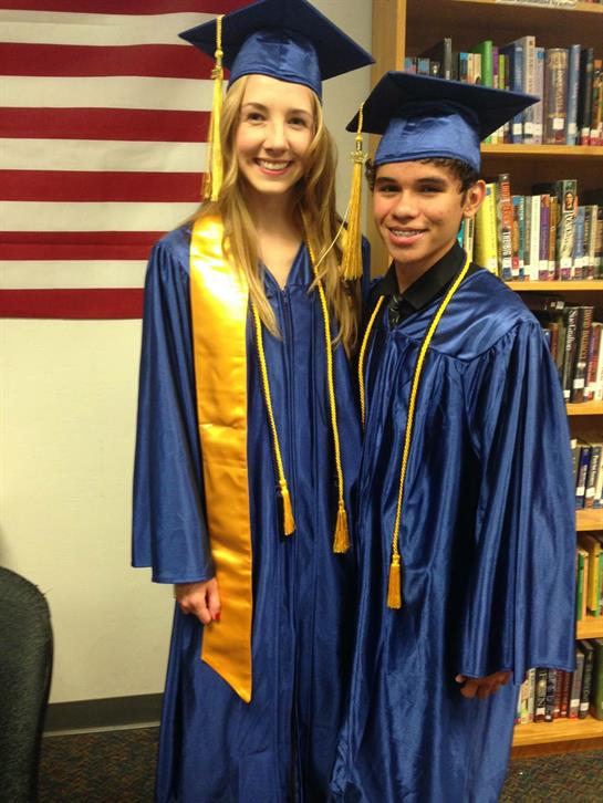 Two graduates proudly pose in blue graduation robes with a background of books and an American flag.