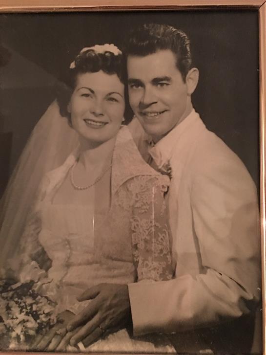 Couple smiles joyfully together in wedding attire, surrounded by floral decorations in a studio.