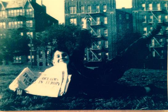 A child smiles while reclining on the grass with a newspaper in hand, surrounded by urban buildings.