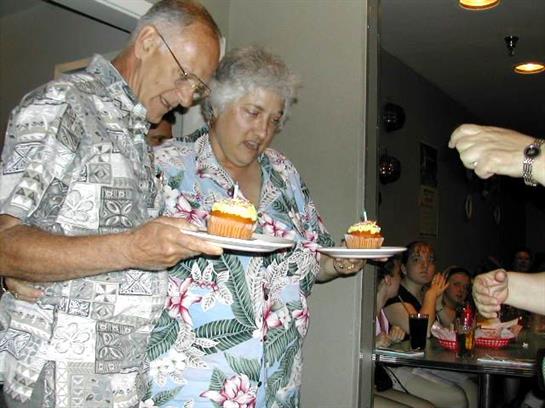 A couple celebrates their anniversary with cupcakes and candles, surrounded by loved ones.