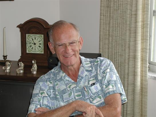 Older man with glasses smiles while seated in a comfortable room with an antique clock nearby.