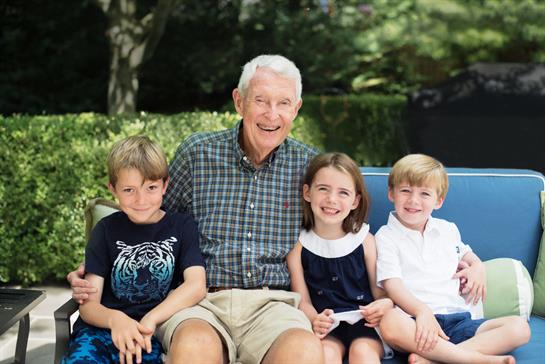 A joyful grandfather sits on a bench with his smiling grandchildren during a sunny afternoon.