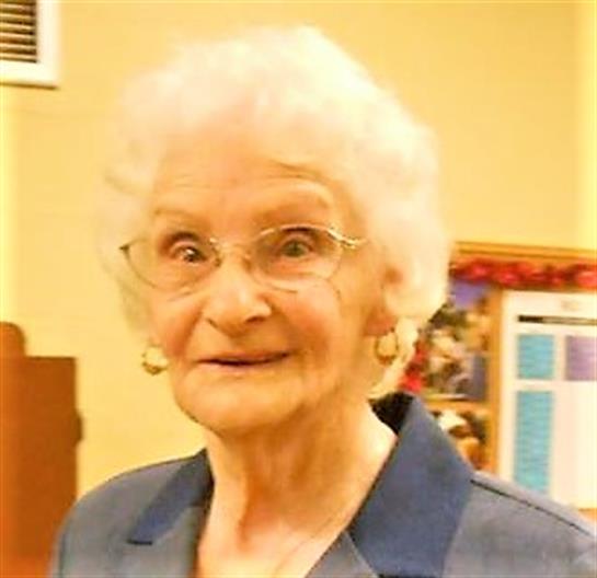 A friendly elderly woman with white hair and glasses smiles joyfully at a community event.