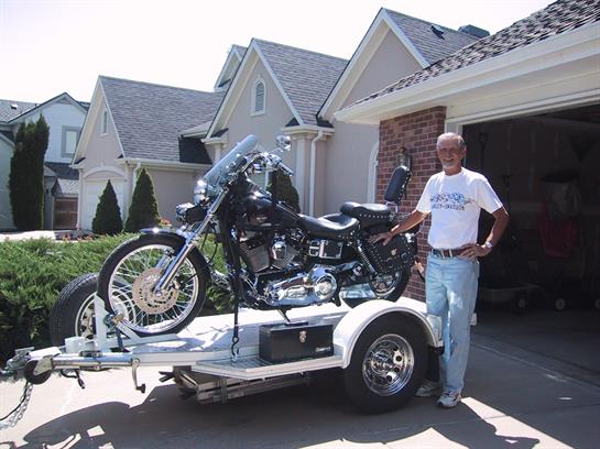 A man poses beside his motorcycle on a trailer in a driveway outside a suburban home.