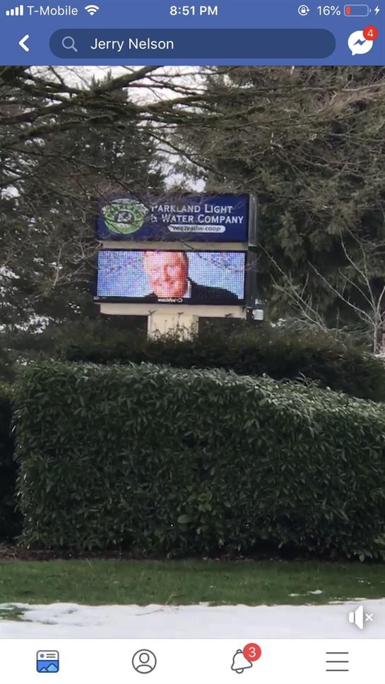 An electronic sign displays a message at Oakland Light and Water Company, amid greenery.