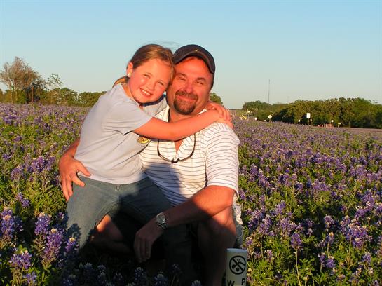 A man and his daughter share a happy embrace in a field of bluebonnets at dusk.