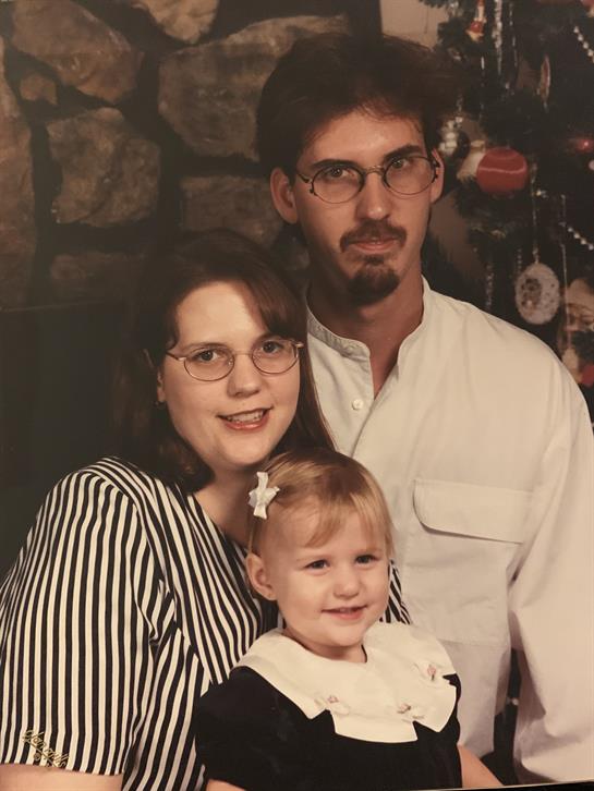 A smiling family poses together in a cozy setting, with mother holding daughter and father nearby.