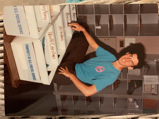 A young man is seated at a workspace surrounded by boxes, smiling while working diligently.