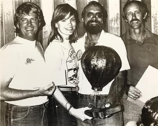 Friends gather casually, showcasing a trophy while dressed in relaxed vintage style.