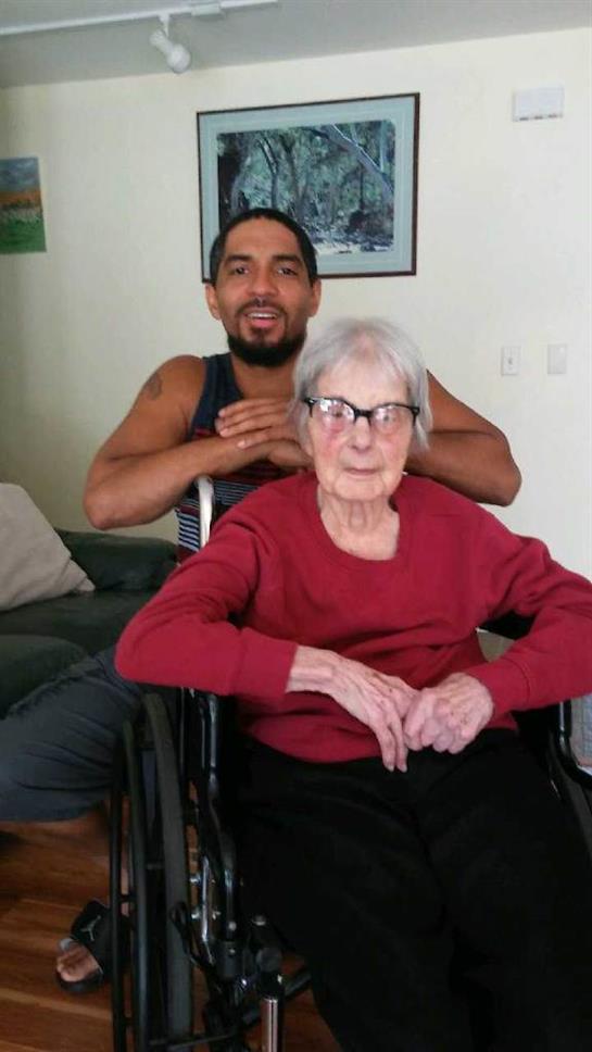 An elderly woman in a wheelchair shares a cheerful moment with a young man in a bright room.
