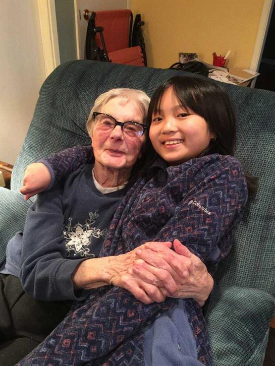 An elderly woman and a young girl smile and embrace on a cozy couch.