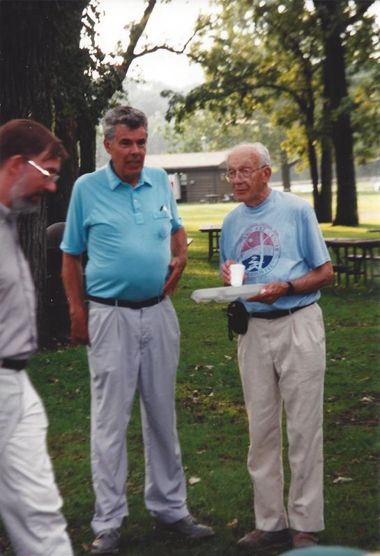 Three men stand together in a park, engaging in conversation while enjoying snacks on a sunny day.