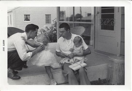 Two men engage with children while seated on a porch on a sunny day.