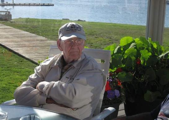 A calm scene shows an elderly man relaxing outdoors by the water on a sunny day.