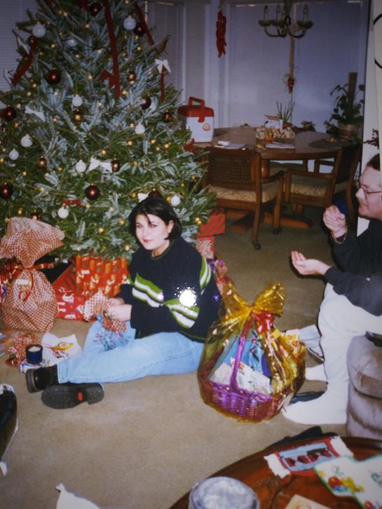 Two people sit on the floor by a decorated tree, surrounded by gifts and festive cheer.