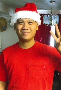 A cheerful young man dressed in a red shirt and Santa hat gives a peace sign while smiling.