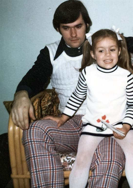 A smiling girl sits on her father's lap, both enjoying a warm and loving indoor moment together.