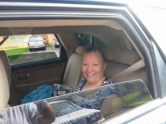 A joyful elderly woman sits inside a car, enjoying a sunny day in a friendly neighborhood.